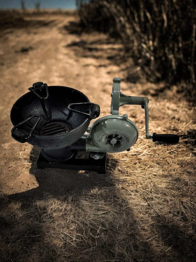 Puede incluir: Un soplador de forja vintage, accionado a mano, pintado en verde oliva y negro. El soplador presenta una carcasa de ventilador circular con el texto "Hazard Factory" y un mango para su funcionamiento. La forja tiene un hogar negro en forma de cuenco.