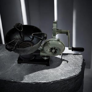 Puede incluir: Un fuelle y un brasero vintage de manivela, probablemente para una fragua. El fuelle es de color verde claro, con un mango negro y una carcasa de ventilador circular. El brasero es negro, con forma de cuenco y tiene un asa. Los objetos est&aacute;n sobre una superficie de piedra.