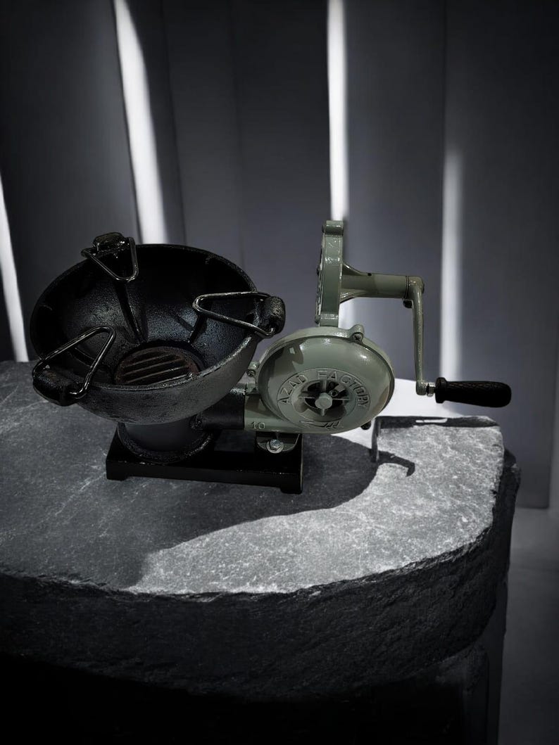 Puede incluir: Un fuelle y un brasero vintage de manivela, probablemente para una fragua. El fuelle es de color verde claro, con un mango negro y una carcasa de ventilador circular. El brasero es negro, con forma de cuenco y tiene un asa. Los objetos est&aacute;n sobre una superficie de piedra.