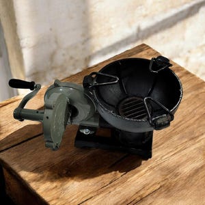 Puede incluir: Una parrilla de carb&oacute;n de manivela de estilo vintage, con un cuenco negro y un componente mec&aacute;nico gris. La parrilla est&aacute; sobre una superficie de madera, mostrando su dise&ntilde;o cl&aacute;sico. El mango es negro.