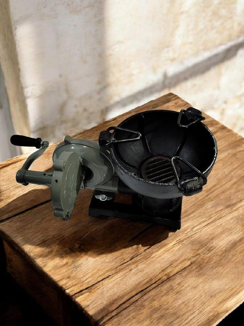 Puede incluir: Una parrilla de carb&oacute;n de manivela de estilo vintage, con un cuenco negro y un componente mec&aacute;nico gris. La parrilla est&aacute; sobre una superficie de madera, mostrando su dise&ntilde;o cl&aacute;sico. El mango es negro.