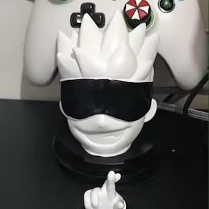 May include: A white figurine of a character with a black blindfold and spiky hair, with a hand gesture in front of it. A white Xbox controller with red and white accents is in the background.