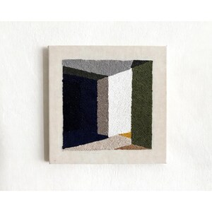 May include: Square, abstract textile art piece. The artwork features a geometric design with a central, open space. The color palette includes shades of gray, navy, white, olive green, and tan. The piece is mounted on a neutral-toned fabric.