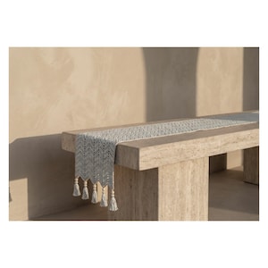 Op de afbeelding: Een lichtgrijze tafelloper met een zigzagpatroon en kwastjes, weergegeven op een stenen bank. De kwastjes van de loper hebben houten kralen. De bank is gemaakt van lichtgekleurde steen en de achtergrond is een neutrale muur.