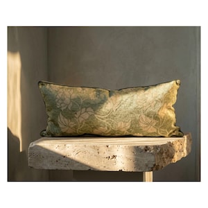 Pode incluir: Uma almofada retangular decorativa com um padrão floral em tons de verde e dourado. A almofada tem uma borda verde escura e é colocada sobre uma superfície de pedra. O design da almofada apresenta um padrão repetido de flores e folhas estilizadas.
