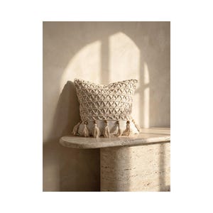 May include: A beige decorative pillow with a geometric pattern and tassels. The pillow is placed on a light-colored stone table. The background is a neutral wall with a shadow.