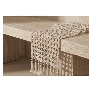 Op de afbeelding: Een crèmekleurige tafelloper met open weefsel, een ruitpatroon en franjekwastjes. De loper is gedrapeerd over een lichtgekleurde stenen tafel. Houten kralen accentueren de kwastjes. De loper voegt textuur en warmte toe.
