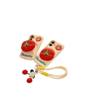 May include: Two off-white phone cases with a 3D red tomato design and green stem. The cases have the word "Tomato" in blue script. A matching wrist strap with a cherry charm is attached.