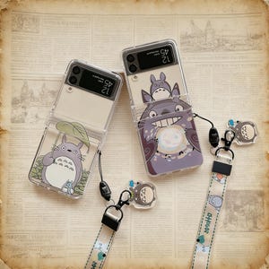 Könnte beinhalten: Zwei transparente Handyhüllen mit Cartoon-Totoro-Designs. Eine Hülle zeigt Totoro unter einem Blatt, die andere zeigt Totoro, der eine leuchtende Kugel hält. Jede Hülle hat ein passendes Schlüsselband mit einem Totoro-Anhänger und dem Text "DANGER".