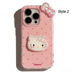 May include: Pink phone case with a Hello Kitty design. The case features a 3D bow and a Hello Kitty face, with small pink polka dots. The word "Happy" is printed at the bottom. The case is designed for a smartphone.