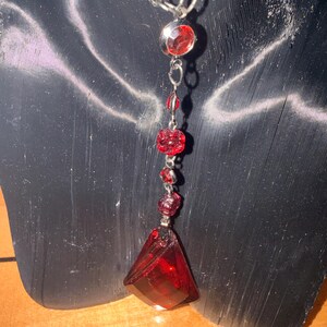May include: A red pendant necklace with a silver chain. The necklace features a series of red, faceted gemstones and a large, triangular pendant. The necklace is displayed on a black mannequin.