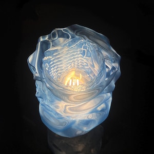 Könnte beinhalten: Eine transluzente blaue Kunstglasskulptur, die einer stilisierten Rose ähnelt. Das Innere zeigt eine warme, leuchtende Lichtquelle, die eine weiche, umgebende Beleuchtung erzeugt. Das Glas hat eine wellenförmige Textur, und das Gesamtdesign ist organisch und fließend.