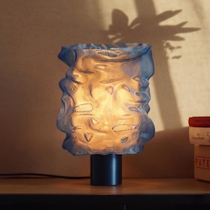Può includere: Una lampada da tavolo blu, strutturata, con base cilindrica. Il paralume ha una forma organica unica e una superficie increspata. La lampada è illuminata, emettendo un bagliore caldo. La lampada è su una superficie di legno.
