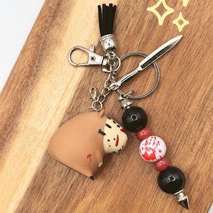 May include: A novelty keychain featuring a brown capybara with a hockey mask design and a small silver spear charm. The keychain includes black and red beads, a black tassel, and a silver clasp.