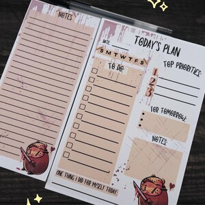 May include: Two notepads with a pen. One notepad has lined paper and the word "NOTES". The other notepad has the words "TODAY'S PLAN", "TOP PRIORITIES", "TO DO", and "FOR TOMORROW". Both notepads have a cartoon character with a knife.
