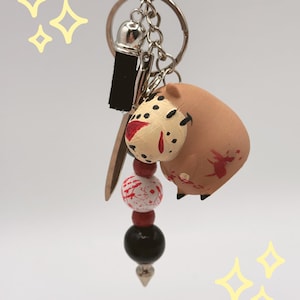 May include: A handmade keychain featuring a brown pig-shaped charm with red blood splatter, a hockey mask, and a small black rectangle. The keychain also includes a series of red, white, and black beads, and a silver-colored metal clasp.