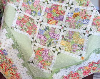 May include: A floral quilt with a green background and a white border. The quilt is made up of squares of different floral fabrics, all with a white background. The quilt is folded in half and is sitting on a brown surface.