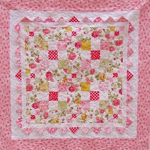 May include: A pink and white quilt with a floral pattern and a checkered border. The quilt is quilted with a diamond pattern.