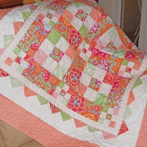 May include: A patchwork quilt featuring a repeating pattern of floral fabric squares in shades of orange, pink, and green, interspersed with white fabric squares. The quilt has a wide border of coral-colored fabric with a small white pattern.