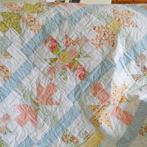 May include: A white quilt with a patchwork design featuring floral patterns in shades of pink, yellow, and green. The quilt is quilted with a diamond pattern.
