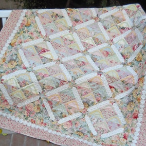 以下が含まれることがあります： ピンク、白、緑のカラーリングのフラワーキルト。キルトはパッチワークの正方形で作られており、縁には白いレースのトリムが施されています。