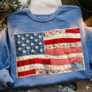 May include: A light blue sweatshirt featuring a patchwork American flag design. The flag includes white stars on a denim blue background, with red and white stripes made of floral and gingham fabrics. The sweatshirt has long sleeves.