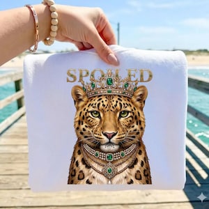 May include: White sweatshirt featuring a leopard illustration wearing a crown and necklace with emerald green accents. The word "SPOILED" is printed above the leopard in gold lettering. The sweatshirt is held up by a person.