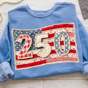 Könnte beinhalten: Ein hellblaues Sweatshirt mit einem patriotischen Design. Das Design zeigt eine amerikanische Flagge im Patchwork-Stil mit den Zahlen "250" aufgenäht. Die Flagge hat rote und weiße Streifen, blaue Sterne und die Zahlen sind in Weiß und Blau.