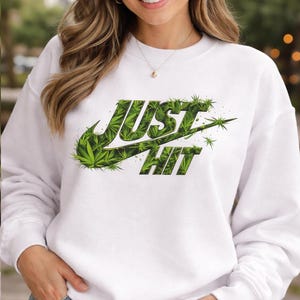 May include: White sweatshirt with the text "JUST HIT" in a green, cannabis-themed design. The design mimics a popular athletic brand logo. The sweatshirt is a casual, comfortable style.