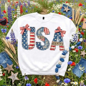 May include: A white long-sleeve sweatshirt featuring the letters USA in a distressed denim and American flag pattern. The letters are accented with red and white striped bows. The design includes patches of leopard print and frayed edges.