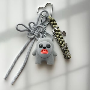 May include: A gray keychain featuring a cartoon character with a red mouth and black eyes. The keychain includes a silver heart-shaped clasp, a gray knotted cord, and a green braided strap with silver charms. The background is white.
