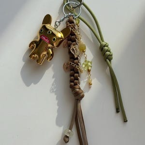 May include: A gold-coloured keychain featuring a stylised figure with multiple eyes and a wide mouth. The keychain includes a braided brown cord, green cord, and decorative charms with leaf and flower accents.