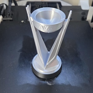 May include: A silver-colored trophy with a circular base and a bowl-shaped top. The trophy has a stylized design with two upward-pointing, V-shaped supports. The top of the trophy has a decorative emblem. The trophy is sitting on a black surface.