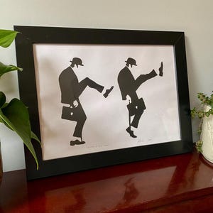 May include: A framed print with a black frame, depicting two black silhouettes of figures in suits and bowler hats, kicking their legs up. Each figure carries a briefcase. The print is signed and has the text "Secrets of Self Magic".