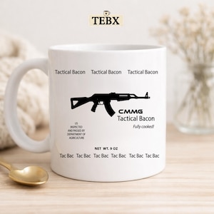 May include: White ceramic mug with the text "Tactical Bacon" and a black graphic of a rifle. The mug also features the text "CMMG Tactical Bacon Fully cooked!" and "NET WT. 9 OZ."