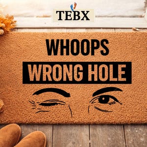 May include: A brown rectangular doormat with the words "WHOOPS WRONG HOLE" in black. Below the text are two cartoon eyes, one winking. The mat is placed on a wooden floor. The brand name "TEBX" is at the top.