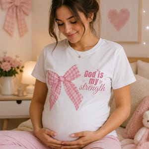 Puede incluir: Una camiseta blanca con un lazo de cuadros rosa y el texto "God is my strength". La camiseta se usa con pantalones rosas. La imagen tiene una estética suave y femenina, con una obra de arte de un corazón rosa en la pared.