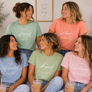 Puede incluir: Cinco camisetas de manga corta y cuello redondo en colores pastel, incluyendo verde salvia, coral, azul claro y rosa claro. Cada camiseta presenta texto en blanco con una referencia bíblica y un nombre personalizado en el pecho.