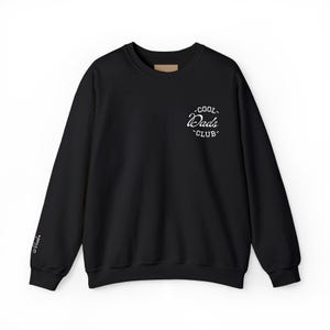 Puede incluir: Una sudadera negra de manga larga con cuello redondo que presenta un gráfico circular blanco en la parte superior izquierda del pecho con el texto COOL DADS CLUB en letras cursivas y de molde. La manga derecha muestra la palabra Noah en letra cursiva blanca.