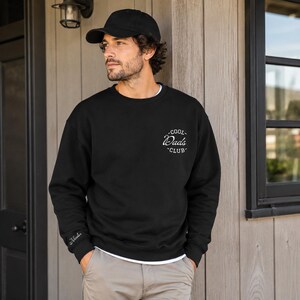 Puede incluir: Una sudadera negra de cuello redondo con un gráfico circular blanco en el pecho izquierdo con el texto Cool Dads Club. El puño de la manga derecha incluye el nombre Nicola bordado en letra cursiva blanca. También se lleva una gorra negra.