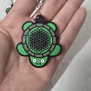 May include: A turtle-shaped keyring featuring a bright green shell with a black geometric flower of life pattern, a black border, and a green head with two white eyes. The turtle is attached to a silver metal split ring by a small chain link.