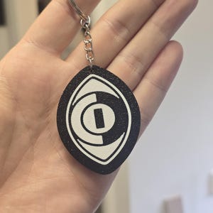 May include: A black and white keychain featuring an almond-shaped pendant with a textured surface. The pendant displays a stylized eye graphic in white against a black background, attached to a silver-toned metal split ring by a short chain.