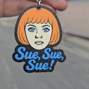 Könnte beinhalten: Ein Schlüsselanhänger mit einer flachen, gestanzten Illustration einer Person mit leuchtend orangefarbenem Haar und blauen Augen. Unter dem Gesicht ist der Text "Sue, Sue, Sue!" in blauen Buchstaben auf schwarzem Grund aufgedruckt. Der Anhänger ist an einer silbernen Metallkette befestigt.