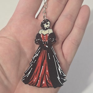 May include: A keyring featuring a flat, illustrated figure wearing a long, formal gown. The dress has a deep red centre panel with black side panels and sleeves, accented by white line details. The figure has dark hair and a pale complexion.