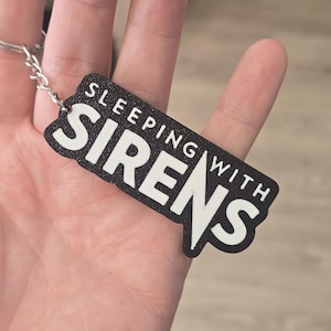 May include: A black rectangular keychain with a textured surface featuring the white text SLEEPING WITH SIRENS in a bold, stylized font. The keychain is attached to a silver-colored metal split ring by a short metal chain link.
