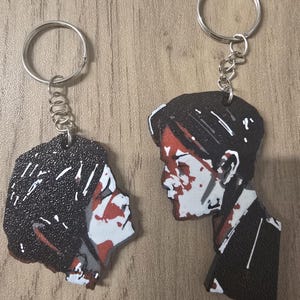 May include: Two keychains featuring stylised illustrations of human profiles. The designs use a high-contrast palette of black, white, and red, depicting faces with dark hair and sharp, angular features. Each keychain includes a silver metal ring.