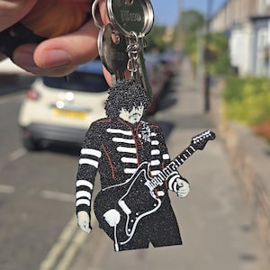 May include: A black glittery keyring featuring a stylised figure with dark curly hair wearing a black military-style jacket with white accents, playing a black electric guitar. The keyring is attached to a metal ring with several silver keys.