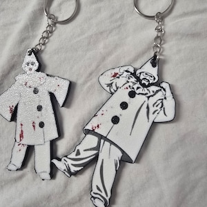 Puede incluir: Dos llaveros con figuras de payasos blancos con botones negros y salpicaduras rojas que parecen sangre. Una figura está de pie y la otra tiene los brazos levantados. Ambos están unidos a anillas de metal plateado con cadenas cortas.