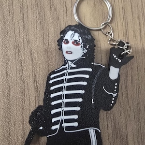 May include: A black and white rubber keychain featuring a figure with dark curly hair, pale skin, and red eye makeup, wearing a black military-style jacket with white piping and a matching glove, attached to a silver metal split ring and chain.