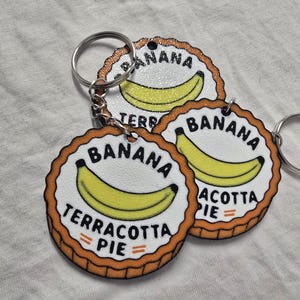 Könnte beinhalten: Drei kreisförmige Schlüsselanhänger mit weißer Mitte, einer gelben Bananenillustration und dem schwarzen Text BANANA TERRACOTTA PIE. Die Ränder sind gewellt und in einem warmen Terrakotta-Orange gehalten, mit einem silbernen Metallring.